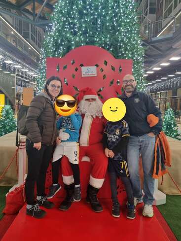 Une famille pose avec le Père Noël devant un grand sapin de Noël décoré dans un centre commercial. Deux enfants ont des autocollants représentant des émoticônes souriantes.