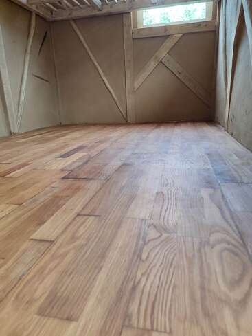 This image shows a wooden floor in a small room with rustic wooden walls and beams. There’s a window at the far end letting in light.