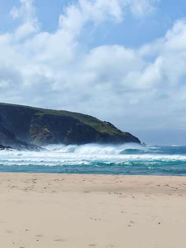 Golden sand stretches across the foreground, leading to turquoise waves crashing against the shore. Lush green cliffs rise behind, beneath a sky of scattered clouds.