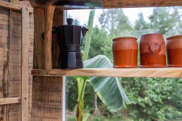 En una estantería de madera hay una cafetera negra y tres tazas de terracota; a través de la ventana que hay detrás se divisa un frondoso jardín y una gran hoja de plátano.