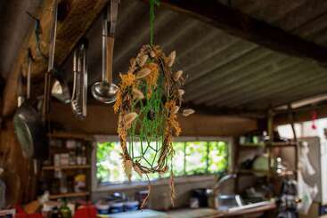 En una cocina rústica, con utensilios de metal, vigas de madera, estanterías abiertas y luz natural, cuelga un atrapasueños hecho a mano con hilo verde, plumas y flores secas.