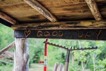 Una estructura rústica de madera con las palabras «GOOD DAY» y un corazón tallado en una viga. Debajo cuelgan herramientas de jardinería, con un fondo de exuberante vegetación.