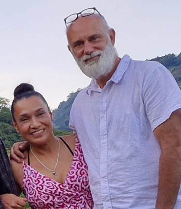 Ein lächelndes Paar steht draußen, mit Bergen im Hintergrund. Der Mann hat weißes Haar, einen Bart und eine Brille auf dem Kopf; die Frau trägt ein rosa Kleid.