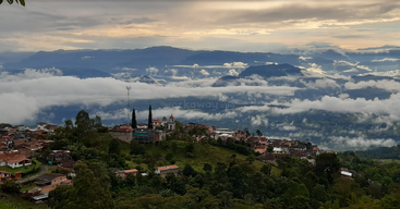 Ein malerisches Bergdorf, umgeben von sattem Grün, Nebel und dramatischen Wolken, mit verstreuten Häusern, einer Kirche und fernen Bergen unter einem leuchtend goldenen Himmel.