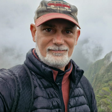Un hombre con barba blanca, que lleva gorra y chaqueta acolchada, sonríe a la cámara frente a un paisaje montañoso y brumoso, probablemente disfrutando de una aventura al aire libre.