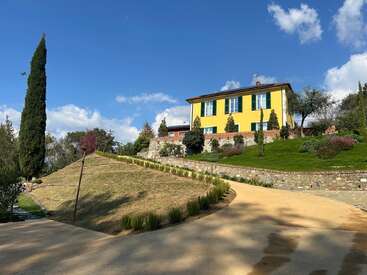Uma casa amarela com persianas verdes fica no topo de uma colina, cercada por vegetação, árvores, um gramado bem cuidado e um céu azul claro com nuvens.