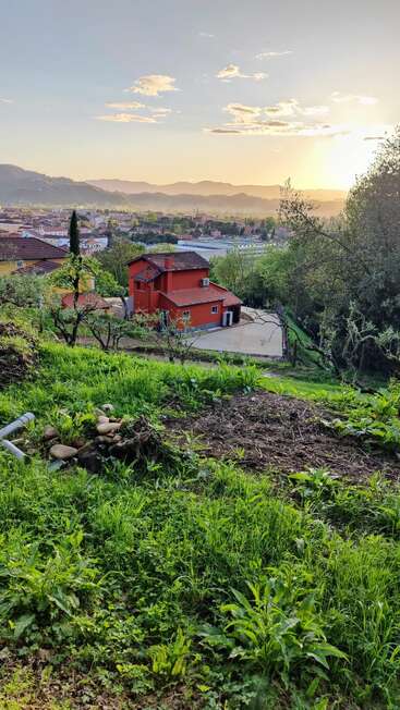 Uma casa laranja vibrante fica em meio a uma vegetação exuberante em uma encosta ao pôr do sol, com vista para uma cidade pacífica com montanhas distantes e um céu brilhante e sereno.