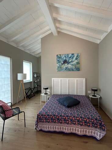 Esta imagem mostra um quarto aconchegante com teto branco inclinado, móveis modernos, uma cama de casal com colcha floral e arte abstrata acima da cama.