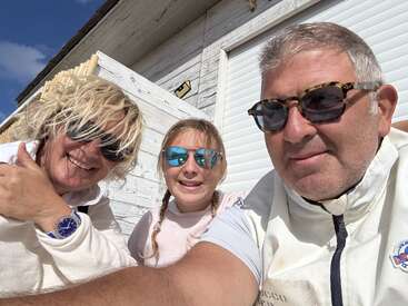 Uma família alegre tira uma selfie ao ar livre. Todos os três usam óculos escuros e roupas de cores claras, sorrindo juntos sob o sol forte ao lado de um prédio de madeira branca.