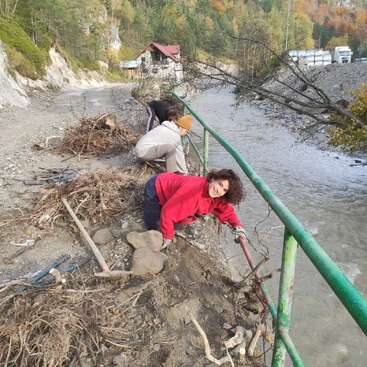 Three people clean debris near a riverbank, clearing branches and rocks by a metal railing. Surrounded by nature, they work together, with one person smiling at the camera.