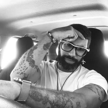Ein bärtiger Mann mit Brille, Tattoos und einer Armbanduhr sitzt in einem Auto. Er wirkt nachdenklich, stützt den Kopf auf die Hand und trägt ein weißes T-Shirt.