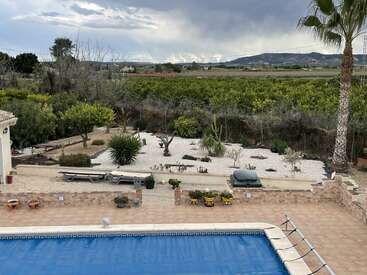 This image shows a backyard with a covered swimming pool, tiled patio, potted plants, lounge chairs, landscaped garden, palm trees, and scenic countryside under a cloudy sky.