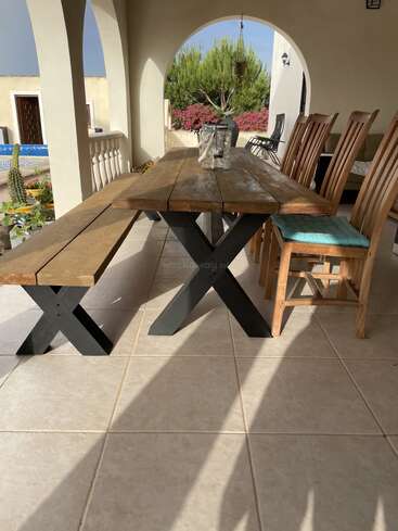 A rustic wooden table with matching bench and wooden chairs sits on a tiled patio. Sunlight streams in, highlighting potted plants, arches, and outdoor scenery.