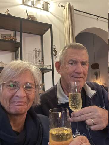 An older couple smiles warmly at the camera, holding glasses of sparkling wine, celebrating together in a cozy, warmly lit indoor setting, surrounded by decor.