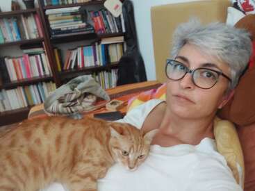 Eine Frau mit kurzen grauen Haaren und Brille entspannt sich auf einer Couch, auf deren Brust eine orangefarbene Katze ruht, umgeben von Bücherregalen.