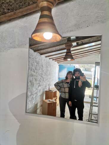 A couple poses together, smiling in front of a mirror, inside a rustic room with exposed beams, wooden furniture, and hanging lights, capturing a cozy moment.