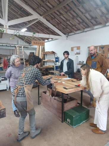 Five people gather around a wooden workbench in a workshop, attentively watching a woodworking demonstration. Tools, wood, and shelves are visible, with a rustic roof overhead.
