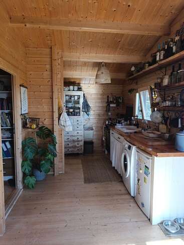 This cozy wooden kitchen features rustic decor, open shelving with jars and bottles, a washing machine, fridge, green plant, woven light fixture, and bright natural lighting.