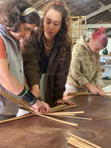 Dans un atelier rustique, trois femmes sont penchées sur une table, occupées à assembler ou à tresser de longs bâtons en bois. Elles semblent très concentrées ; elles participent peut-être à un cours d'artisanat ou de bricolage.