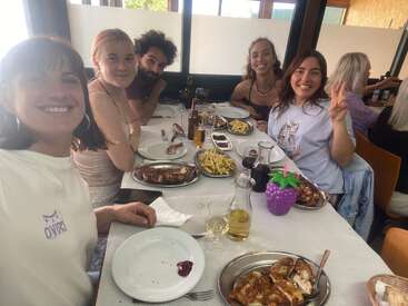 Cinq amis sourient à une table de restaurant, en train de savourer un repas composé de poulet rôti et de frites. L'un d'eux fait le signe de la paix, créant une ambiance joyeuse.