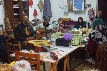 Un groupe de personnes est réuni à l'intérieur autour d'une table, occupé à faire du bricolage et de la musique. Certains jouent de la guitare tandis que d'autres tricotent, dessinent et discutent dans une ambiance chaleureuse.