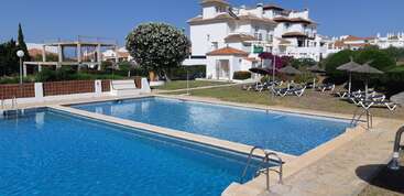 A clear blue swimming pool with sun loungers and straw umbrellas, surrounded by green grass and white buildings under a bright, sunny sky. Relaxing atmosphere.