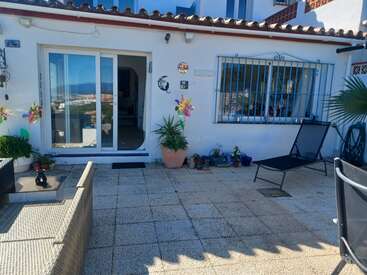 A sunny patio with tiled flooring, lounge chairs, potted plants, pinwheel decorations, and a sliding glass door. Blue metal window bars and outdoor furniture complete the cozy space.