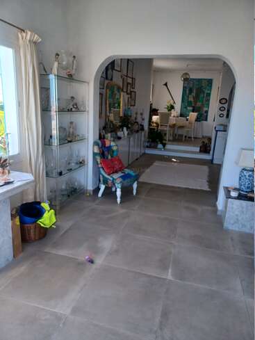 A bright, cozy living space with large tiles, glass display cabinet, vibrant armchair, basket, and connecting archway leading to a decorated dining area with art.