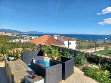 A sunny terrace with modern patio furniture overlooks a scenic coastline, colorful houses, lush gardens, blue sea, distant hills, and bright, clear skies above.