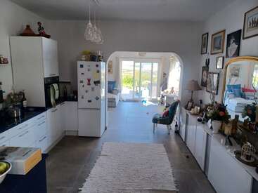 Bright and cozy kitchen-living room with white cabinets, fridge, art-filled walls, mirror, indoor plants, comfortable seating, and patio doors opening to a sunlit garden.