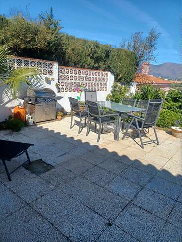 A sunny patio with a glass dining table, four chairs, barbecue grill, decorative wall, green bushes, and clear blue sky, perfect for outdoor gatherings.
