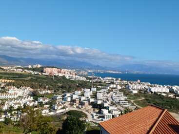 This image shows a coastal town with white buildings, red-tiled roofs, lush green hills, and a blue sea under a bright, clear sky with clouds.