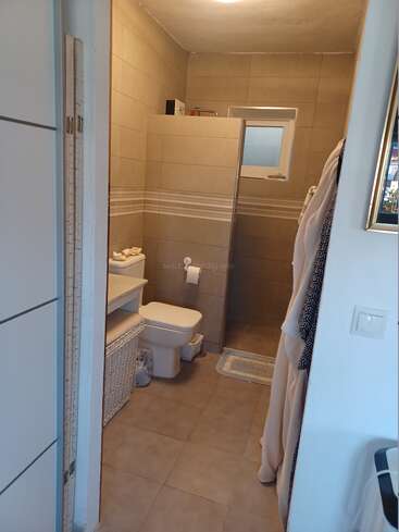 This image shows a modern bathroom with beige tiles, a toilet, a shelf with towels, a wicker basket, a shower area, and a small window.