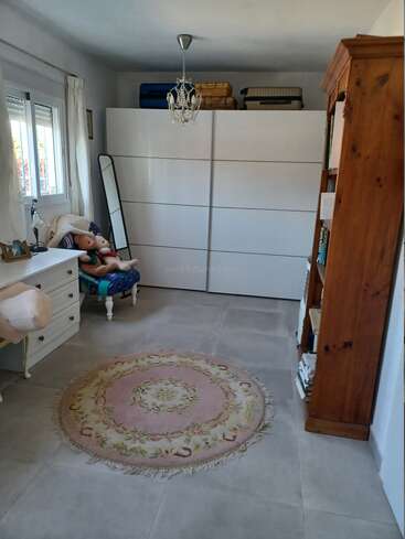 A cozy room with a white wardrobe, wooden bookshelf, round pink rug, dresser, window, mirror, chandelier, armchair with stuffed animals, and suitcases on top.