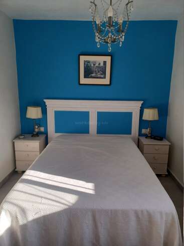 A cozy bedroom with a white bed, blue accent wall, two bedside tables, matching lamps, a framed picture above the headboard, and a decorative chandelier. Sunlight streams in.