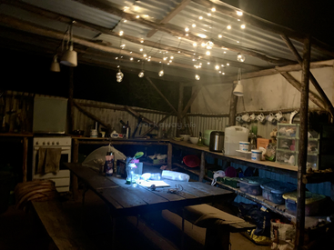 Eine gemütliche, rustikale Außenküche mit Holzbänken und -tischen, von der Decke hängenden Lichterketten, Regalen mit Vorräten und einem Ofen in der Ecke.