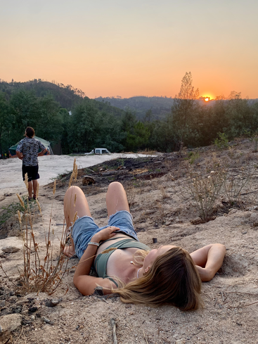 Eine Frau liegt entspannt auf dem Sandboden und blickt in den Sonnenuntergang. Ein Mann steht in der Nähe, umgeben von Bäumen und Hügeln, und genießt die friedliche, natürliche Landschaft.