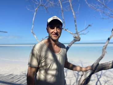 A man stands by a tree on a bright sunny beach, wearing a cap and beige t-shirt. The ocean and blue sky create a serene background.