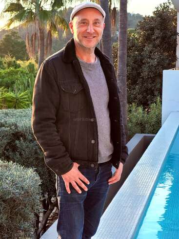 Ein Mann, der einen grauen Pullover, eine schwarze Jacke und eine weiße Mütze trägt, steht an einem Pool und lächelt. Palmen und üppiges Grün bilden den malerischen Hintergrund.