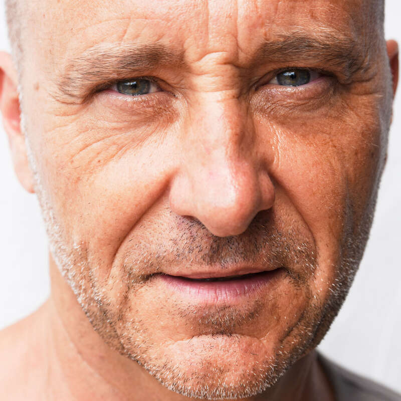 Gros plan sur le visage d'un homme d'âge moyen, montrant des yeux bleus, une barbe courte, des rides, une peau bronzée et une expression sérieuse et réfléchie. L'arrière-plan est neutre.