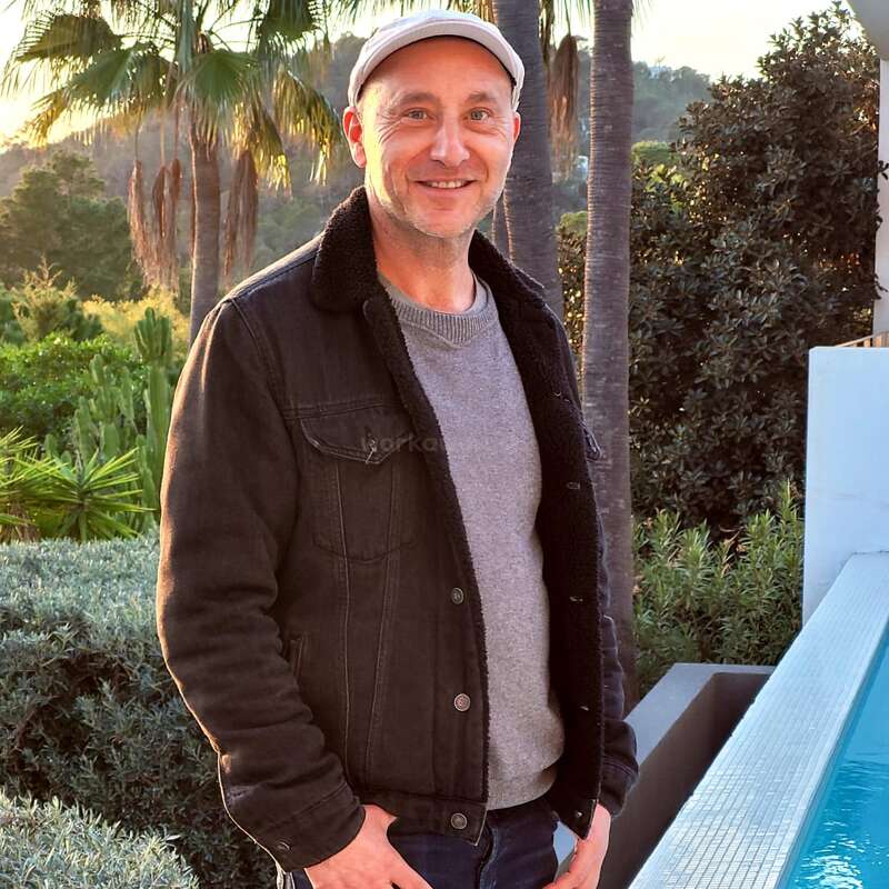 A man wearing a gray sweater, black jacket, and white cap stands by a pool, smiling. Palm trees and lush greenery form the scenic background.