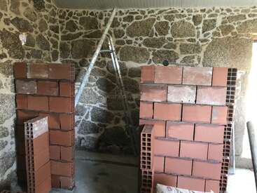 Una habitación en construcción presenta un muro de piedra, dos pilas de ladrillos rojos, una escalera metálica apoyada en la pared y materiales de construcción esparcidos por los alrededores.