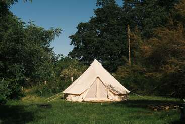 Una gran tienda de lona de color crema está montada sobre la hierba verde, rodeada de frondosos árboles y arbustos, bajo un cielo azul y despejado. Una tranquila escena de acampada.