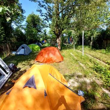 Several colorful tents are set up in a grassy, shaded area surrounded by trees under a bright blue sky. The scene gives a peaceful, outdoor camping vibe.