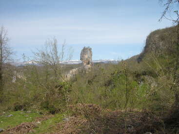 A solitary tall rock formation rises dramatically from a lush green valley, surrounded by trees and distant hills beneath a clear blue sky with mountains beyond.