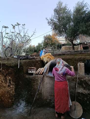 A woman in traditional clothing is baking bread in an outdoor clay oven, surrounded by trees and rural scenery, with tools and loaves nearby, creating a rustic atmosphere.
