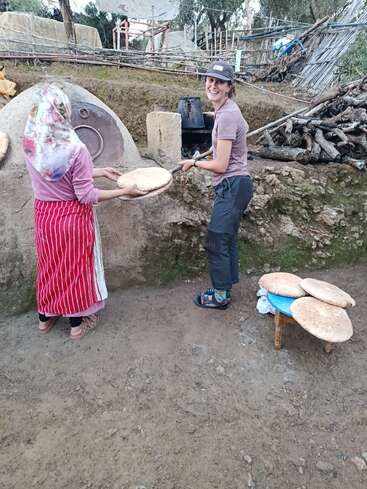 Two women are baking traditional bread outdoors beside a clay oven. One woman is handing over bread dough, while the other smiles, holding a baking tool.