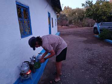 A person in casual clothes prepares tea outdoors, using a kettle and stove beside a white building with blue windows, surrounded by trees and a parked vehicle.