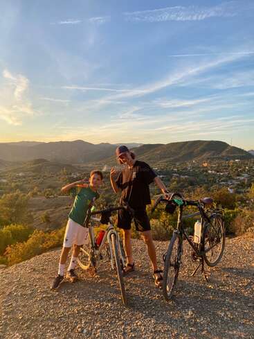 Two people pose happily with their bicycles on a hilltop, surrounded by scenic mountains, greenery, and a beautiful blue sky during sunset, enjoying outdoor adventure.