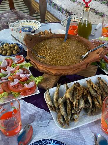 A traditional meal is served, featuring a large bowl of lentil soup, fried fish, fresh tomato salad, olives, bread, olive oil, and colorful drinks.